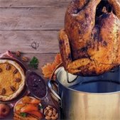 Fried turkey being lifted from a large pot with Thanksgiving foods like pie, vegetables, and nuts arranged on a wooden table.