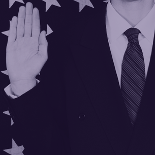 close up photo of man in black suit with right hand raised and us flag in background
