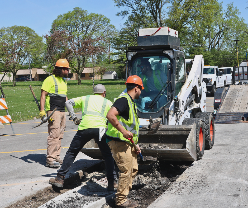 Public Works crew repairing pavement with a skid steer and hand tools in a parking lot